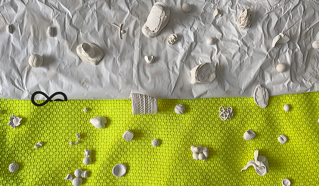 An image description for this image is: The image is separated by a horizontal line. The upper part of the picture is made of white paper and the lower one is made of neon yellow fabric with knitted structure. On the on both backgrounds are scattered various small marbles, stones and clay shapes. A curved infinity neurodiversity sign connects both backgrounds.